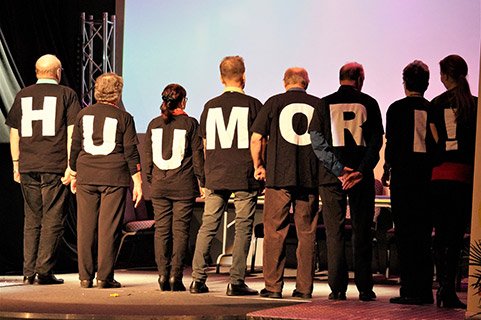 Lavalla seisomassa henkilöitä selin yleisöön päin: paitojen selkäosassa on kirjaimia, joista tulee sana huumori